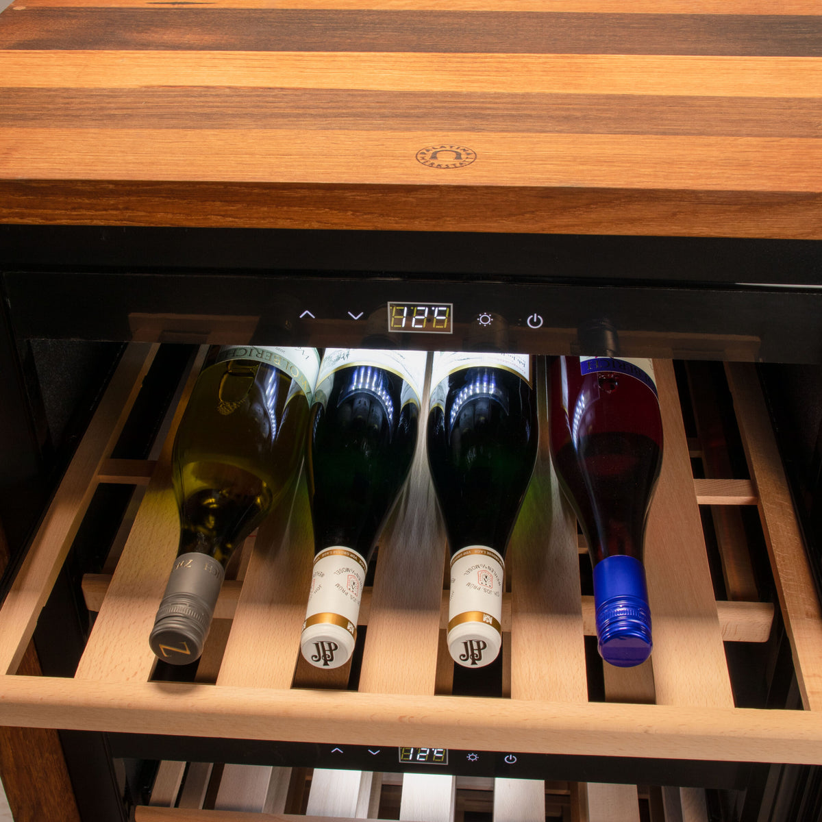 Eine Nahaufnahme des 2-Zonen-Weinkühlschranks CASO WineExclusive 66 Smart in schwarz mit Fassholz-Umhausung zeigt eine auf 12 °C eingestellte Digitalanzeige. Der Kühlschrank verfügt über ein Holzregal mit Platz für vier Flaschen Wein, darunter Weiß- und Roséweinsorten, mit beschrifteten Flaschenhälsen und einer Mischung aus Schraub- und Korkverschlüssen. Für zusätzlichen Komfort ist er per APP steuerbar.
