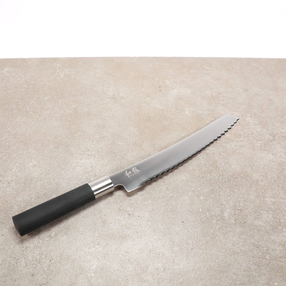 Ein Kai Wasabi Black Brotmesser mit 23 cm Klinge und schwarzem Griff liegt auf einem großen Schneidebrett aus Eiche, 40 x 30 cm groß. Die gezackte Klinge hat ein silbernes Finish und weist ein eingraviertes Symbol in der Nähe des Griffs auf, das elegant auf der strukturierten grauen Oberfläche ruht.