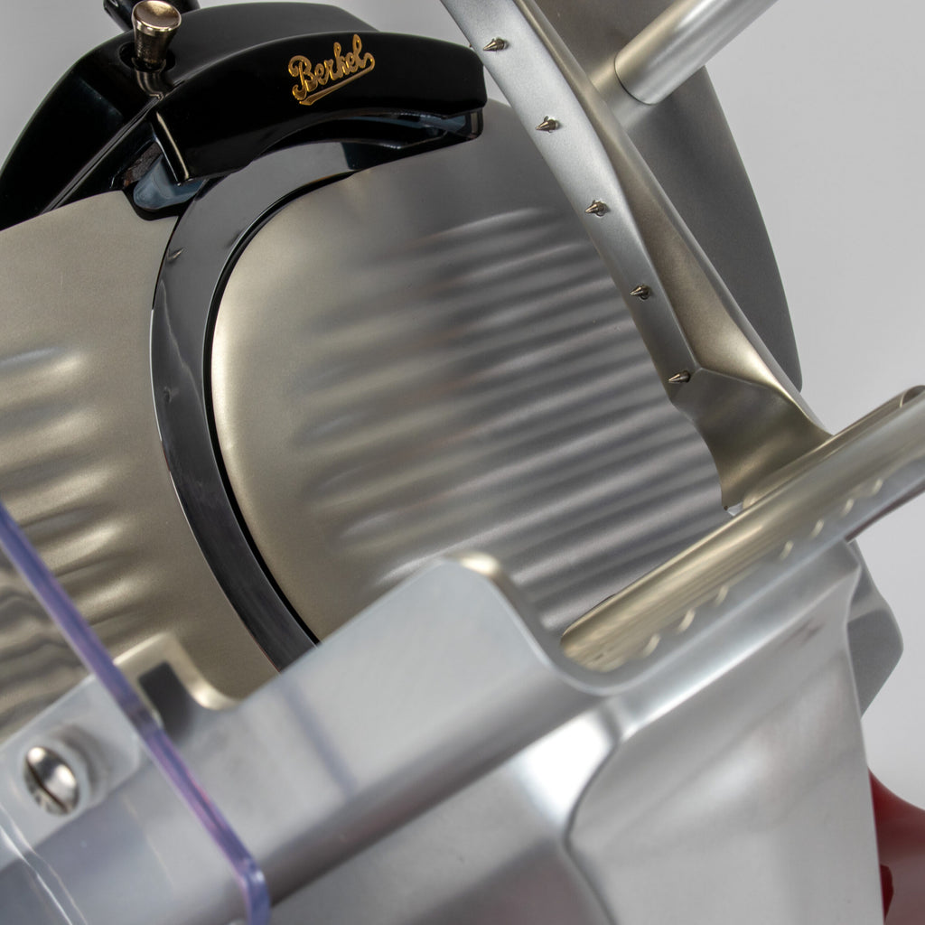 Nahaufnahme einer Berkel Aufschnittmaschine Pro Line XS 25 Rot, Der Profi Allesschneider für Ihre Küche + 100 % handgefertigtes Fassholzbrett Unikat, mit gezahnter Klinge und metallischem Finish. Der Allesschneider ist mit einem Griff zum Einstellen der Schnittstärke ausgestattet. Das Gesamtdesign wirkt langlebig und für den professionellen Einsatz gedacht.