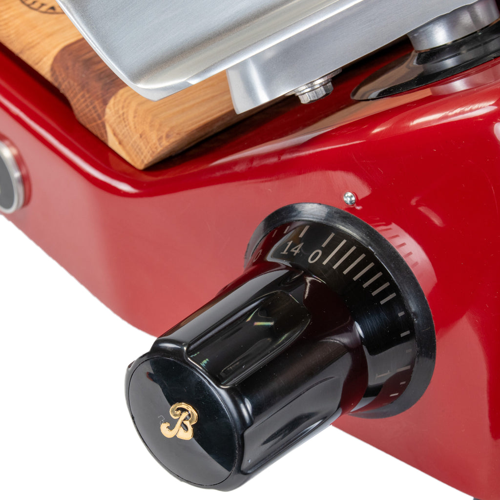 Nahaufnahme einer Berkel Aufschnittmaschine Pro Line XS 25 Rot, mit Fokus auf einem schwarz-goldenen Drehknopf zum Einstellen der Schnittstärke. Der Drehknopf ist auf Stufe 14 eingestellt und trägt das Markenlogo „B“. Im Hintergrund sind auch Holz- und Metallteile dieses Profi-Allesschneiders zu sehen, ergänzt durch ein 100 % handgefertigtes Fassholzbrett-Unikat.