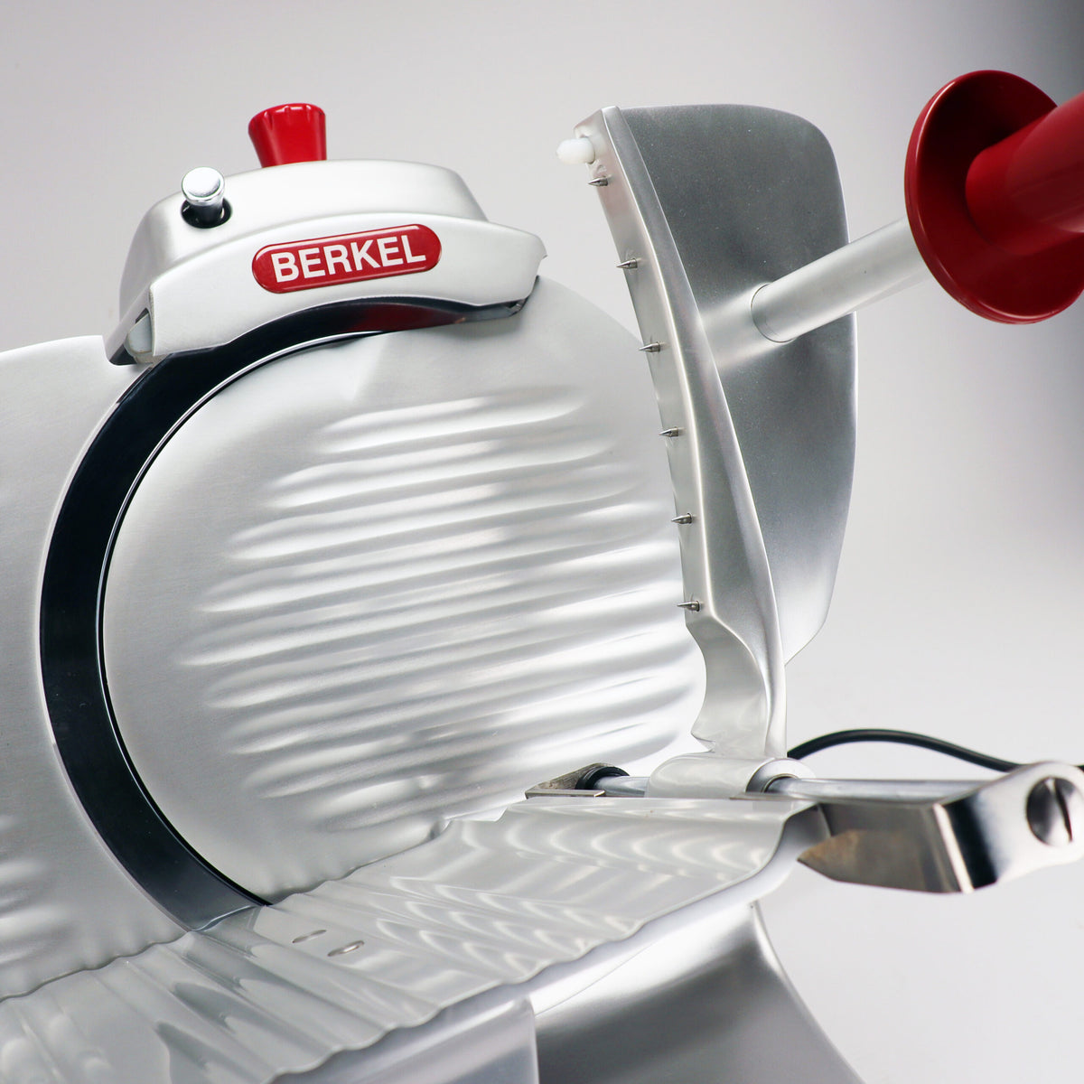 Bild einer Berkel Pro Line XS 25 Silber, professionelle Aufschnittmaschine + Fassholz-Einlegebrett mit rotem Knopf und Griff, mit der Aufschrift „Berkel“ auf einem rot-weißen Etikett. Der Allesschneider verfügt über eine runde, gezahnte Klinge und eine Edelstahlplattform zum Halten des Fleisches. Der Hintergrund ist hellgrau.
