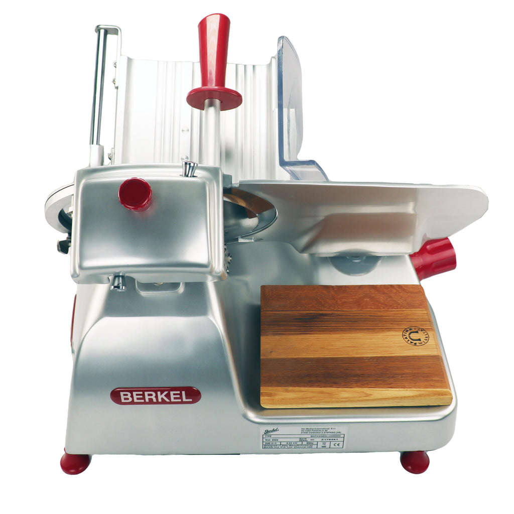 Die Berkel Pro Line XS 25 Silber, professionelle Aufschnittmaschine + Fassholz-Einlegebrett ist eine Fleischschneidemaschine aus Metall mit roten Akzenten, auf deren Vorderseite der Markenname „Berkel“ deutlich zu sehen ist. Sie ist mit einem Klingenschutz, einem einstellbaren Drehknopf für die Schnittstärke und einer hölzernen Schneideplattform auf der rechten Seite ausgestattet, die mit einem runden Logo verziert ist.