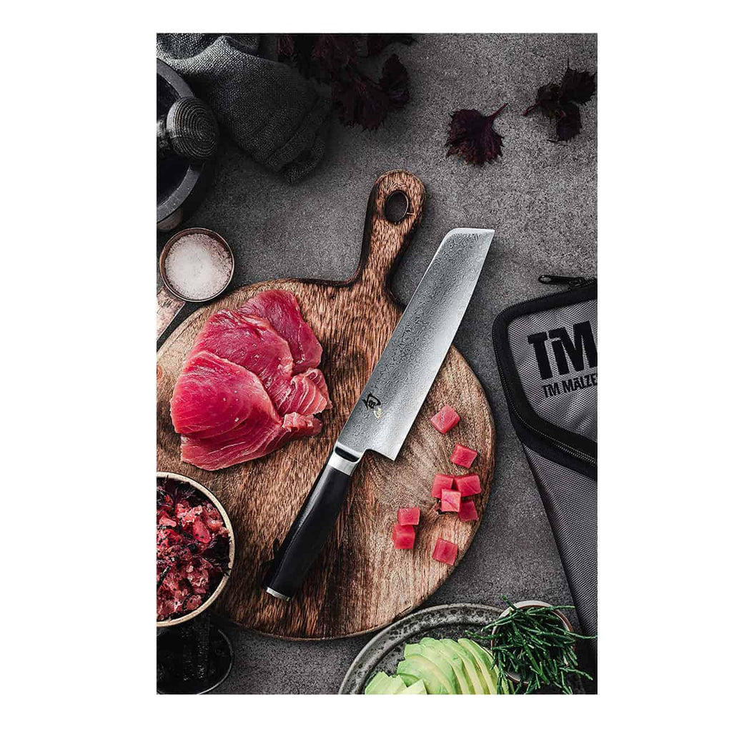 Ein Kai Shun Premier Tim Mälzer TMM-0702 Minamo großes Santokumesser 18 cm auf einem handgefertigten Schneidebrett aus Fassholz 30x18cm mit geschnittenem rohem Thunfisch. Um das Brett herum stehen kleine Schüsseln mit Gewürzen, gewürfelten Zutaten, einer Avocado und einem großen Santokumesseretui von Tim Mälzer auf einer grauen Arbeitsplatte.