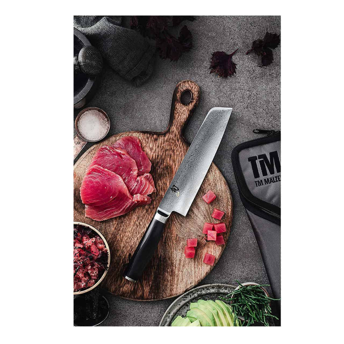 Ein Kai Shun Premier Tim Mälzer TMM-0702 Minamo großes Santokumesser 18 cm auf einem handgefertigten Schneidebrett aus Fassholz 30x18cm mit geschnittenem rohem Thunfisch. Um das Brett herum stehen kleine Schüsseln mit Gewürzen, gewürfelten Zutaten, einer Avocado und einem großen Santokumesseretui von Tim Mälzer auf einer grauen Arbeitsplatte.