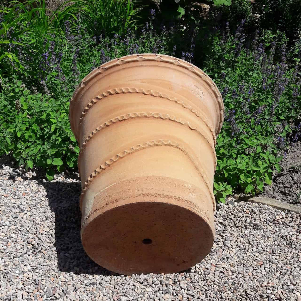 Ein großer Terrakotta-Pflanzkübel „Acanthus“, 50 cm hoch, liegt auf der Seite zwischen grünem Laub und kleinen, violetten Acanthus-Blüten. Der Topf weist ein dekoratives Spiralmuster auf und steht auf einer Kiesfläche in einer Gartenumgebung.