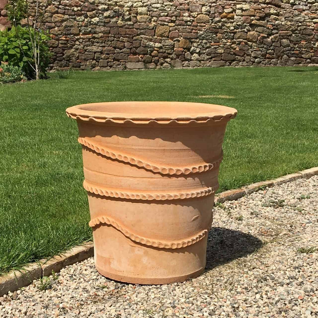 Ein großer Terrakotta-Pflanzkübel „Acanthus“, H=50 cm, mit dekorativen Wellenlinien und gezackten Kanten steht auf einem Kiesweg. Im Hintergrund sind ein gepflegter Rasen, eine Backsteinmauer und etwas Grün zu sehen.
