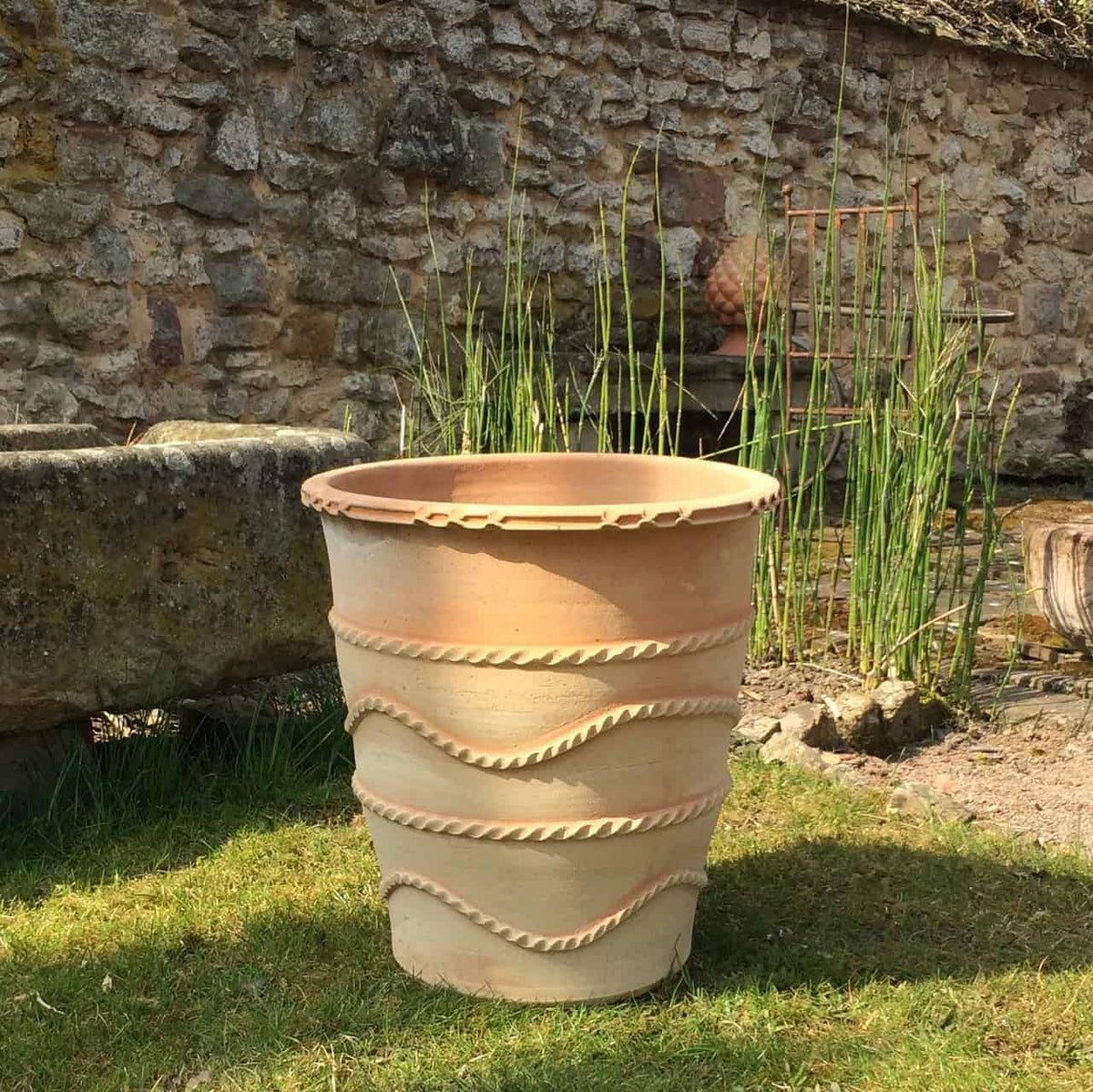 Ein 50 cm hoher, beigefarbener Terrakotta-Pflanzkübel „Acanthus“ mit dekorativen gewellten und gedrehten Mustern steht auf dem Rasen. Dahinter befindet sich eine Steinmauer und einige hohe, dünne Grünpflanzen. Die Szene scheint in einem Garten oder Außenbereich an einem sonnigen Tag zu sein, wobei die Acanthus-Blüten der Umgebung Charme verleihen.