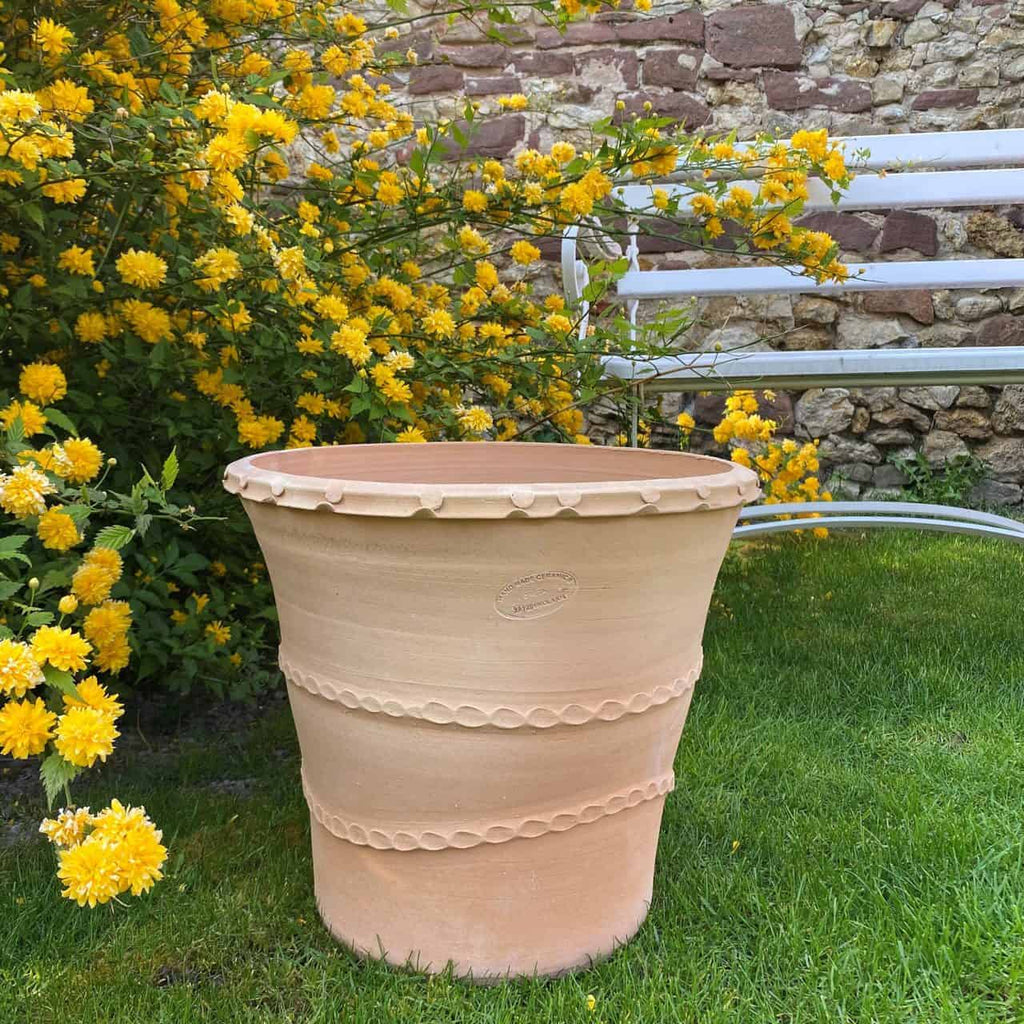 Ein großer, leerer Terrakotta-Pflanzkübel „Acanthus“ mit einer Höhe von 40 cm steht auf grünem Gras neben einer weißen Metallbank. Hinter Topf und Bank steht ein Busch mit leuchtend gelben Blüten in voller Blüte, vor einer rustikalen Steinmauer, die mit Acanthusblättern geschmückt ist.