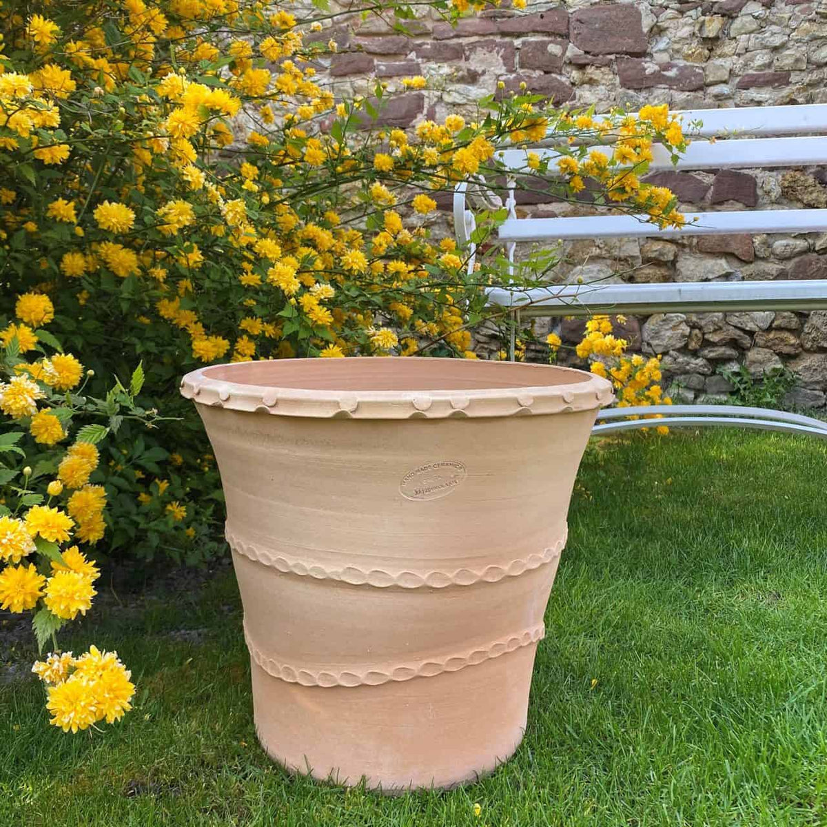 Ein großer, leerer Terrakotta-Pflanzkübel „Acanthus“ mit einer Höhe von 40 cm steht auf grünem Gras neben einer weißen Metallbank. Hinter Topf und Bank steht ein Busch mit leuchtend gelben Blüten in voller Blüte, vor einer rustikalen Steinmauer, die mit Acanthusblättern geschmückt ist.