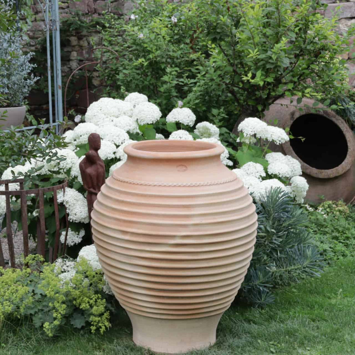 Ein großer, beigefarbener, gerippter Keramiktopf steht in einem Garten, umgeben von weiß blühenden Pflanzen und Grünpflanzen. Neben dem Topf steht eine kleine, braune Statue, und im Hintergrund liegt eine weitere frostfeste Terrakotta-Amphore „Agave“ auf der Seite. Frostfeste Agavenpflanzen zieren die üppige und ruhige Szene.