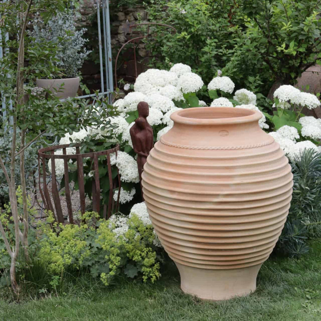 Eine große, frostfeste Terrakotta-Amphore „Agave“, 100 cm hoch, ist umgeben von blühenden weißen Hortensien und üppigem grünem Laub in einem Garten. Eine kleine Figur steht neben der Amphore, während im Hintergrund teilweise eine Metallstruktur sichtbar ist. Eine Agavenpflanze verleiht der ruhigen und grünen Szene eine markante Note.