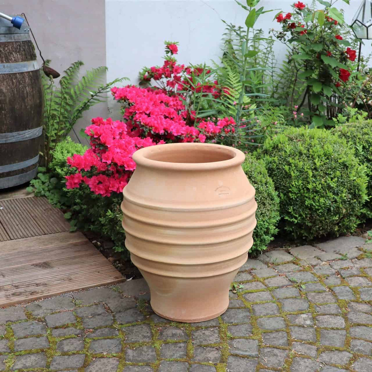 Eine große, gerippte, frostfeste Dekoamphore „Agave“ (H=50cm) steht auf einer gepflasterten Terrasse. Der Pflanzkübel ist leer und steht neben leuchtend rosa Blumen, grünen Sträuchern und einem Holzfass. Im Hintergrund sind Teile einer Holzterrasse und eine weiße Wand mit Kletterpflanzen zu sehen.