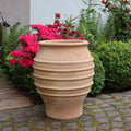 Ein großer, beiger, gerippter Keramiktopf steht auf einer mit Steinen gepflasterten Fläche neben einem Garten. Um die frostfeste Dekorationsamphore „Agave“, H=50cm herum befinden sich leuchtend rosa Blüten, dichte grüne Sträucher und üppiges Blattwerk, die einen farbenfrohen und kontrastreichen Hintergrund bilden.