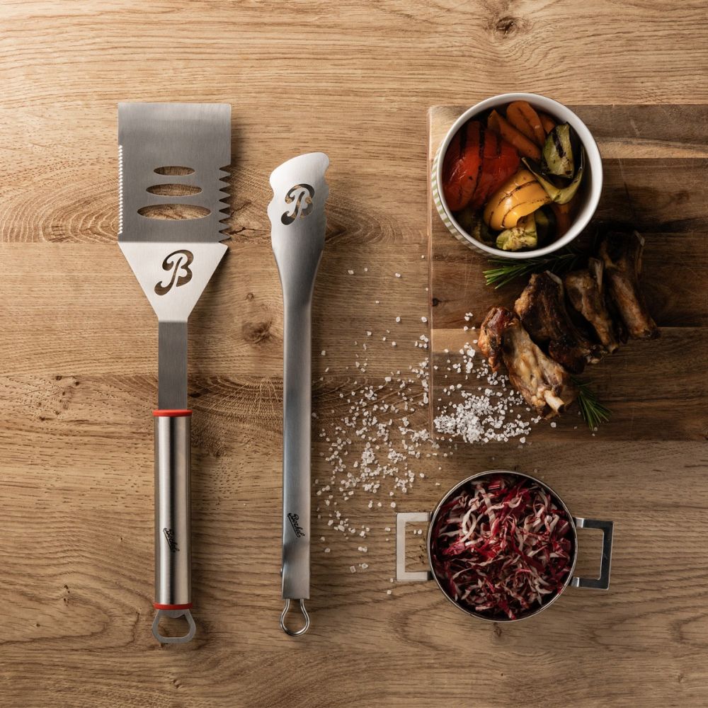 Das Berkel BBQ-Set mit Spachtel und Zange liegt auf einer Holzplatte neben einem Schneidebrett mit gegrilltem Gemüse, BBQ-Ribs, Salz und einem kleinen Topf mit zerkleinertem Rotkohl.