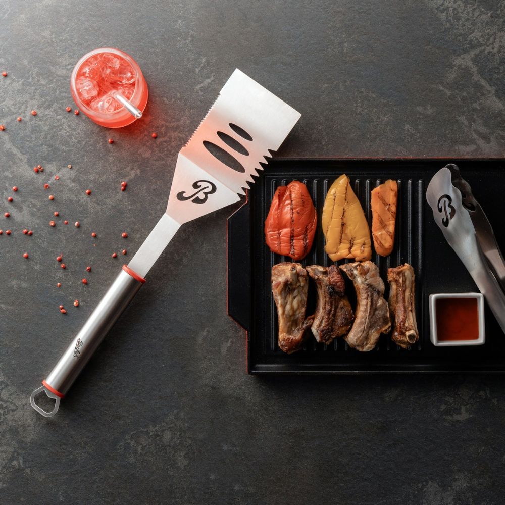 Das Berkel BBQ-Set besteht aus einer Grillschale mit Fleisch und Gemüse, einer Metallzange und einem Spatel mit den Initialen "B" auf einer dunklen Oberfläche. Daneben steht ein rotes eisgekühltes Getränk mit aufgestreuten roten Pfefferkörnern.