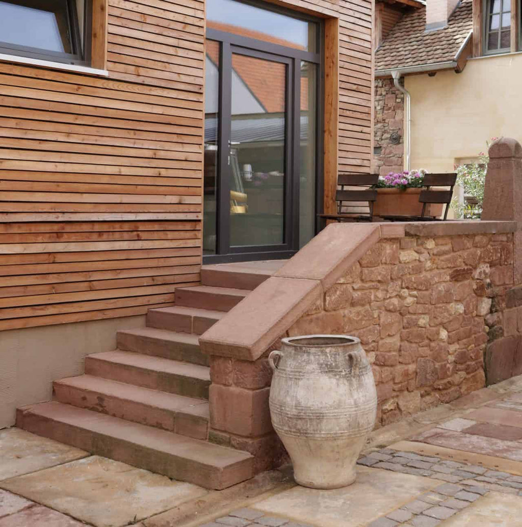 Ein rustikaler Terrassenbereich verfügt über Steinstufen, die zu einer Tür mit großen Glasplatten führen. Die Wände sind mit horizontalen Holzlatten verkleidet. Am Fuß der Stufen steht eine große Antiquarische Amphore 4. Oben auf den Stufen steht ein kleiner Tisch mit Topfblumen.