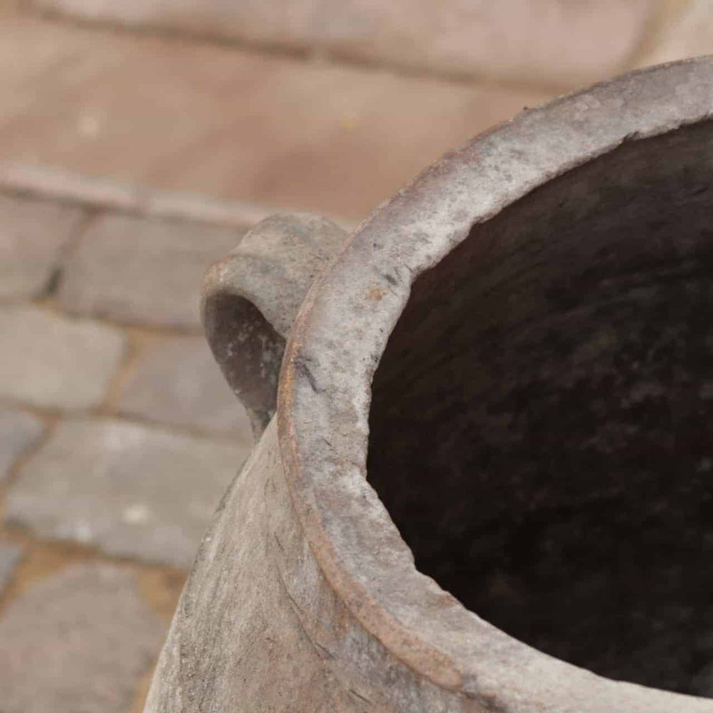 Abgebildet ist ein verwitterter Keramiktopf mit Henkel, identifiziert als Antiquarische Amphore 4. Der Rand des Gefäßes wirkt rau und erodiert. Der Hintergrund ist aus Steinpflaster.
