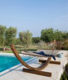 Eine ruhige Szene im Freien mit einer hölzernen Hängematte komplett mit Netzwerk neben einem Pool. Über der Hängematte hängt eine Decke, und üppige grüne Bäume und Sträucher vervollständigen den Hintergrund. Der Himmel ist klar und blau und trägt zur ruhigen Atmosphäre bei.