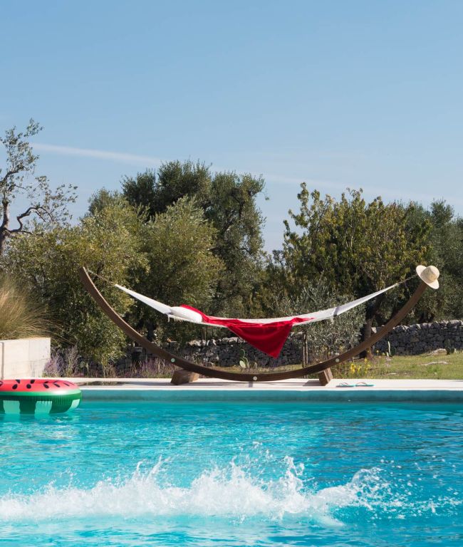 In einer ruhigen Szene im Freien ziert ein Swimmingpool den Vordergrund, während im Hintergrund eine Hängematte komplett mit Netzwerk zu sehen ist. Diese Hängematte, geschmückt mit einer roten Decke und einem breitkrempigen Hut darauf, liegt zwischen Bäumen unter einem klaren blauen Himmel. Die idyllische Kulisse wird noch durch eine schwimmende Wassermelone im Pool unterstrichen.
