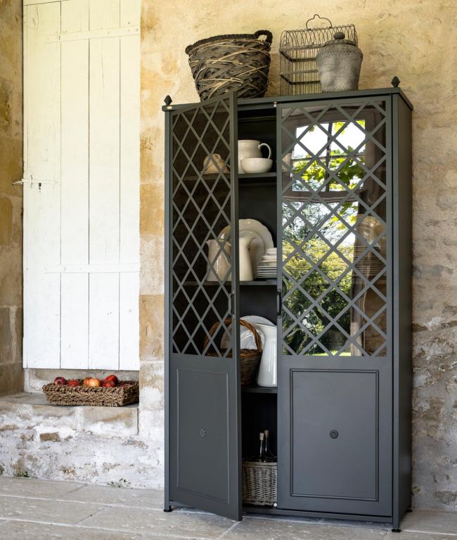 Ein hoher Schrank Aurora aus Metall mit gitterförmigen Glastüren steht vor einer Steinwand. Die teilweise geöffneten Türen geben den Blick auf Regale frei, die mit verschiedenen Gerichten und Dekorationsgegenständen gefüllt sind. Auf und neben dem Schrank stehen geflochtene Körbe mit Äpfeln und anderen Gegenständen.
