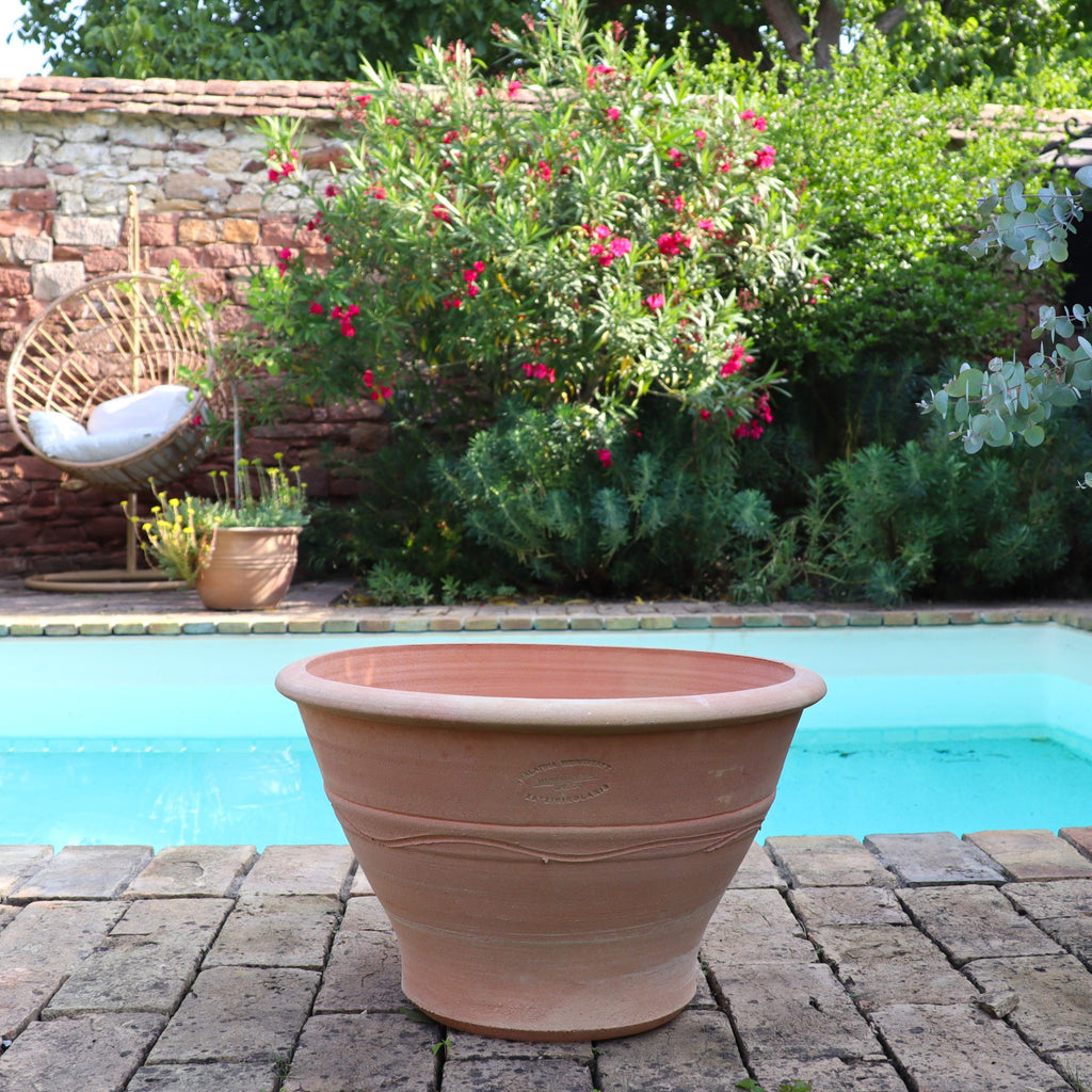 Ein geräumiger Terrakotta-Pflanztopf "Artemesia", B=45cm, steht leer auf einem Ziegelsteinpflaster neben einem klaren blauen Teich, umrahmt von einem üppigen grünen Garten, einem rosa blühenden Busch und einem hängenden Gartenstuhl auf der linken Seite.