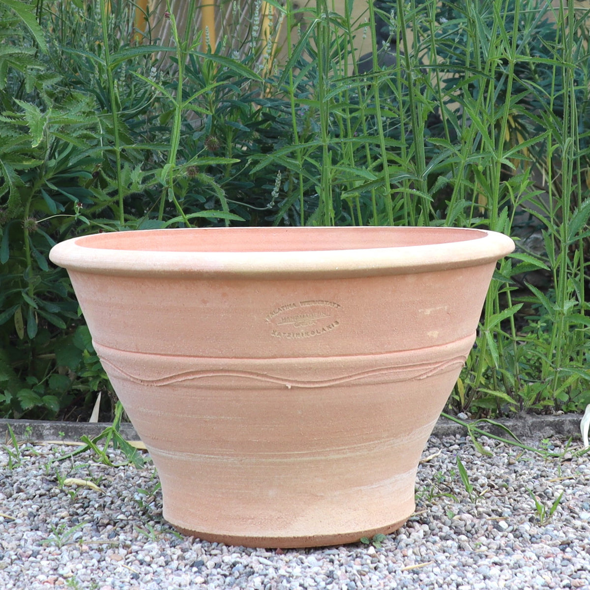 Der Terracotta-Pflanztopf "Artemesia", B=45cm steht leer auf Kies, umgeben von hohen Grünpflanzen und Gras.