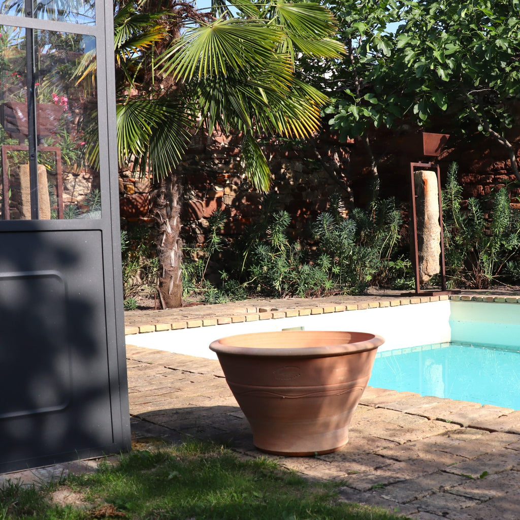 Der Terrakotta-Pflanztopf "Artemesia", B=45cm, steht leer auf einer gepflasterten Terrasse an einem blauen Pool, umgeben von Palmen und üppigem Grün. Das Sonnenlicht wirft weiche Schatten auf die ruhige Umgebung.