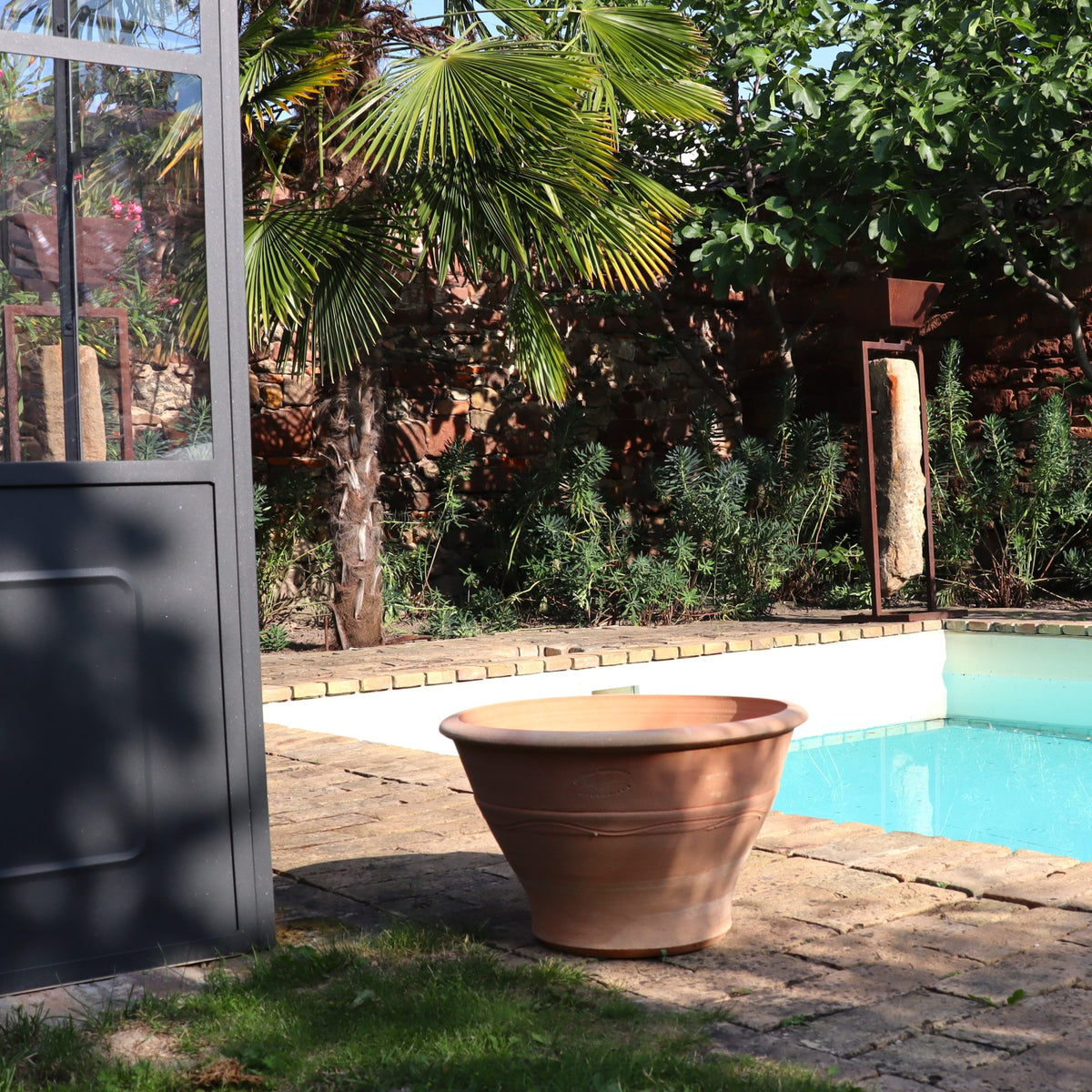 Der Terrakotta-Pflanztopf "Artemesia", B=45cm, steht leer auf einer gepflasterten Terrasse an einem blauen Pool, umgeben von Palmen und üppigem Grün. Das Sonnenlicht wirft weiche Schatten auf die ruhige Umgebung.