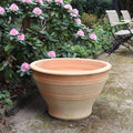 Ein großer, leerer Terrakotta-Pflanzkübel steht auf einem Gartenweg neben einem Busch mit rosa Blüten. Im Hintergrund sind ein Gartenstuhl und ein Holztisch zu sehen. Die Szene ist hell und ruhig und suggeriert eine ruhige Gartenumgebung mit einem eleganten winterfesten großen Pflanzkübel, Blumenkübel 70 cm, Artemesia, der den Charme noch verstärkt.
