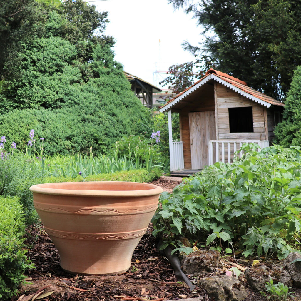 Ein winterfester großer Pflanzkübel steht auf einem gemulchten Gartenweg, umgeben von üppigem Grün. Im Hintergrund ist ein kleiner Holzschuppen mit weißem Geländer zu sehen, eingerahmt von verschiedenen Bäumen und Sträuchern. Links vom Weg blühen violette Blumen, dazwischen sind Stränge von Artemisia eingestreut, die für zusätzliche Struktur sorgen.