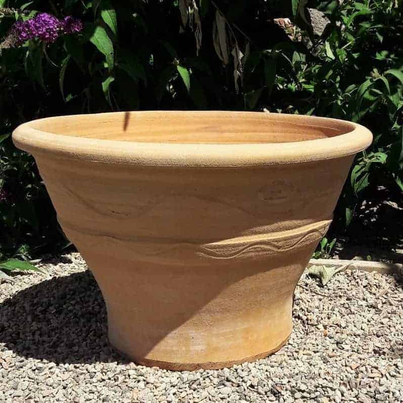 Ein großer, hellbrauner Terrakotta-Pflanztopf „Artemisia“ mit einer Breite von 50 cm steht auf einem Kiesbett. Der Pflanztopf hat eine breite, ausgestellte Öffnung und ist in der Mitte mit einem einfachen, geprägten Wellenmuster verziert. Im Hintergrund sind üppige grüne Pflanzen mit einigen violetten Blüten, darunter Artemisia, zu sehen.