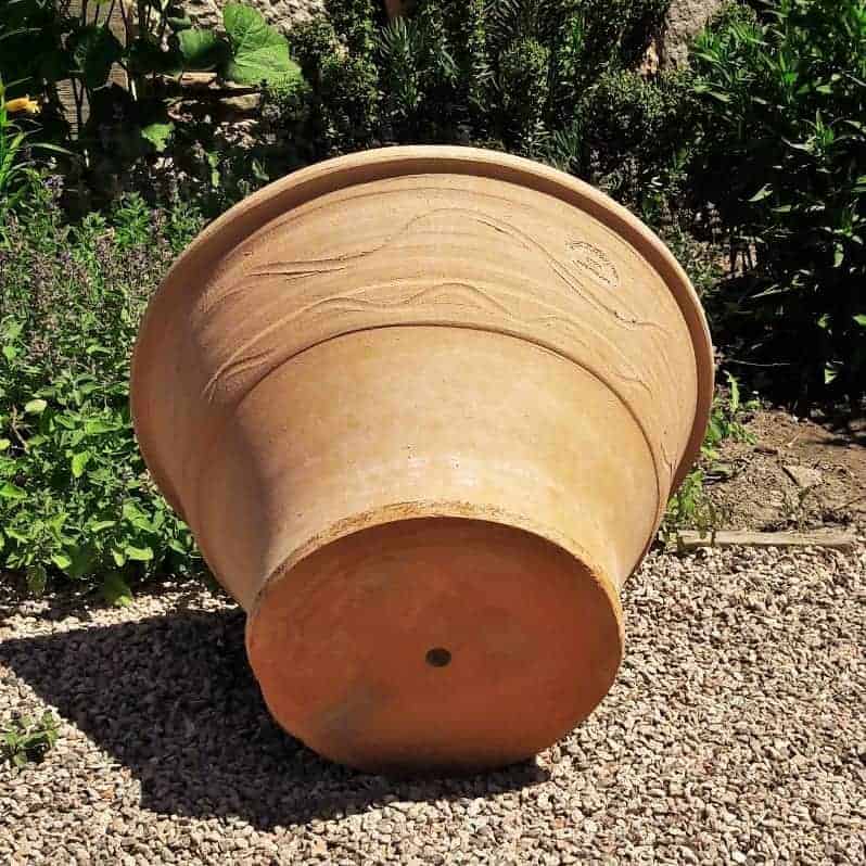 Ein großer Terrakotta-Pflanztopf „Artemisia“ mit aufwendigem Wellenmuster liegt seitlich auf einem Kiesboden. Der 50 cm große Topf steht in einer Gartenecke, umgeben von verschiedenen Grünpflanzen und Sträuchern.