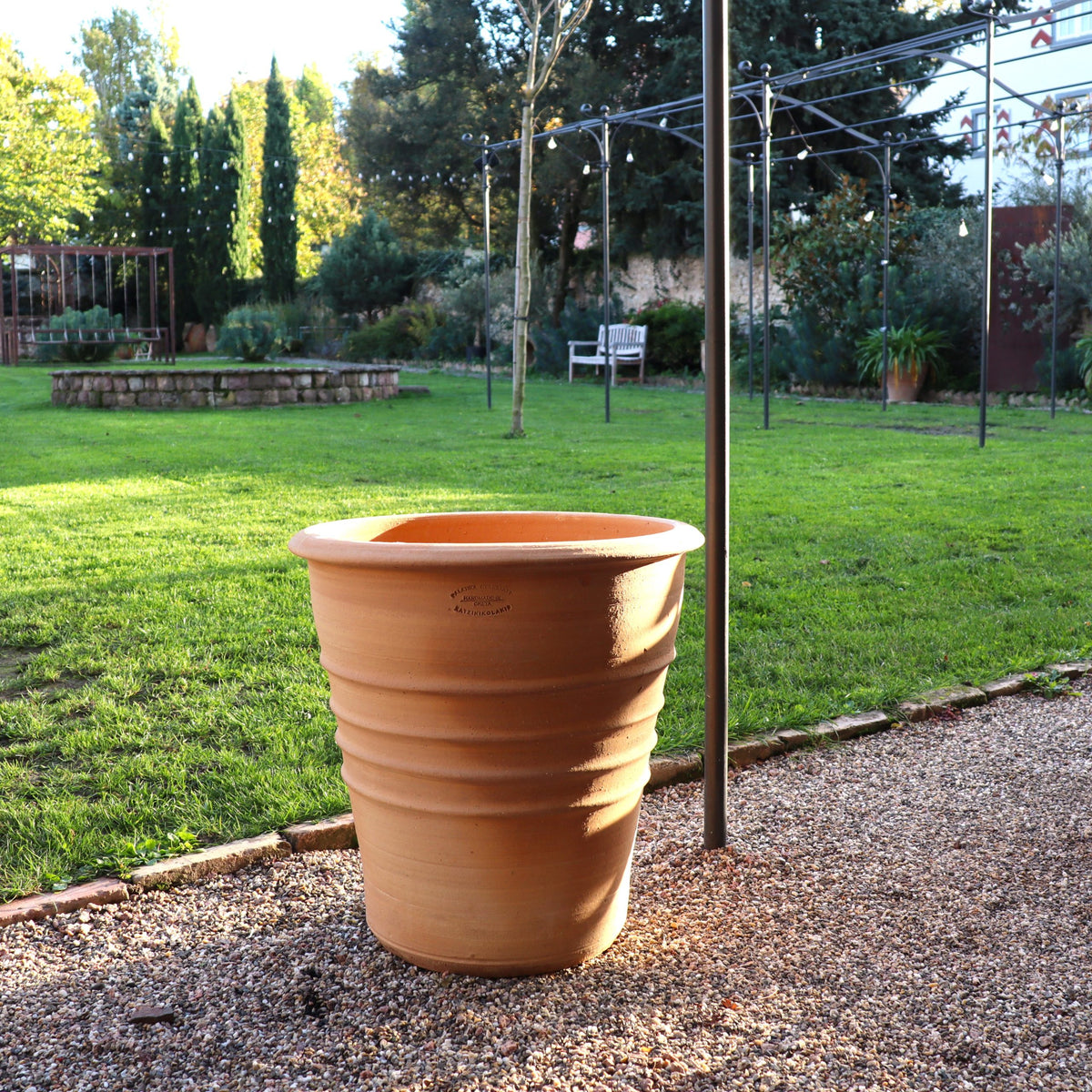 In einem hellen Garten steht der Terracotta Blumentopf "Aster", H=50 cm, leer auf Kies neben einem grünen Rasen mit Bäumen, einer Bank und Lichterketten.