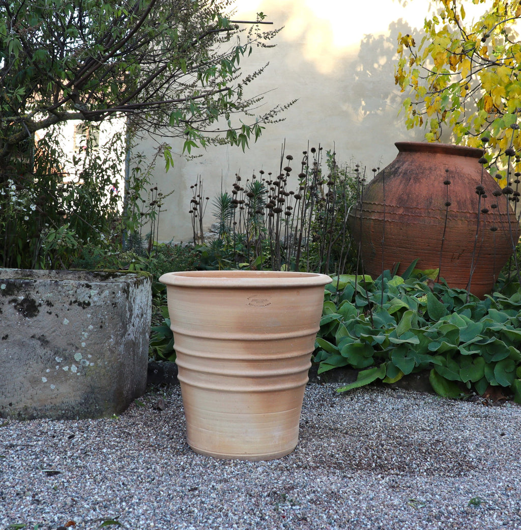 Der Terracotta Blumentopf "Aster", H=50 cm, in Hellbraun, steht auf Kies in einem Garten mit Grünpflanzen, einem steinernen Übertopf und einem weiteren Tontopf an einer beigen Wand.