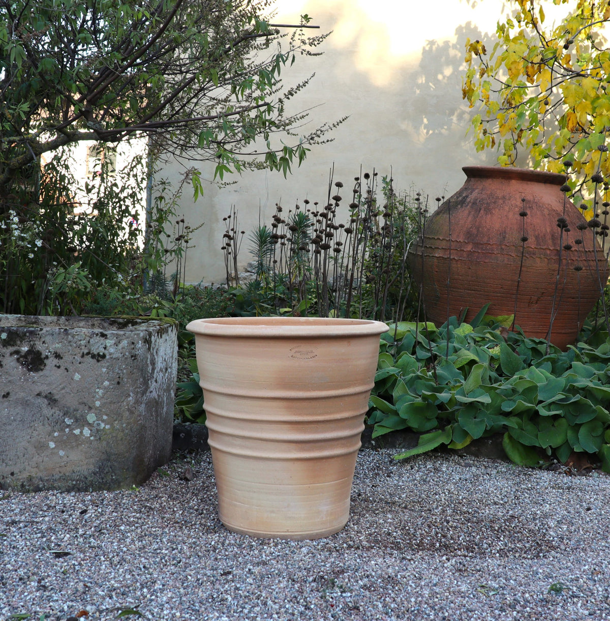 Der Terracotta Blumentopf "Aster", H=50 cm, in Hellbraun, steht auf Kies in einem Garten mit Grünpflanzen, einem steinernen Übertopf und einem weiteren Tontopf an einer beigen Wand.