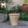 Der Terrakotta-Blumentopf "Aster" (H=50 cm) steht auf einem Kiesweg im Garten, umrahmt von üppigem Blattwerk, mit einem steinernen Pflanzgefäß und einem großen Tonkrug im Hintergrund.