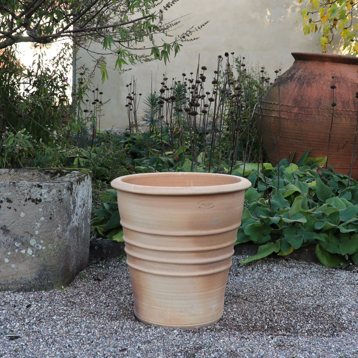 Der Terrakotta-Blumentopf "Aster" (H=50 cm) steht auf einem Kiesweg im Garten, umrahmt von üppigem Blattwerk, mit einem steinernen Pflanzgefäß und einem großen Tonkrug im Hintergrund.