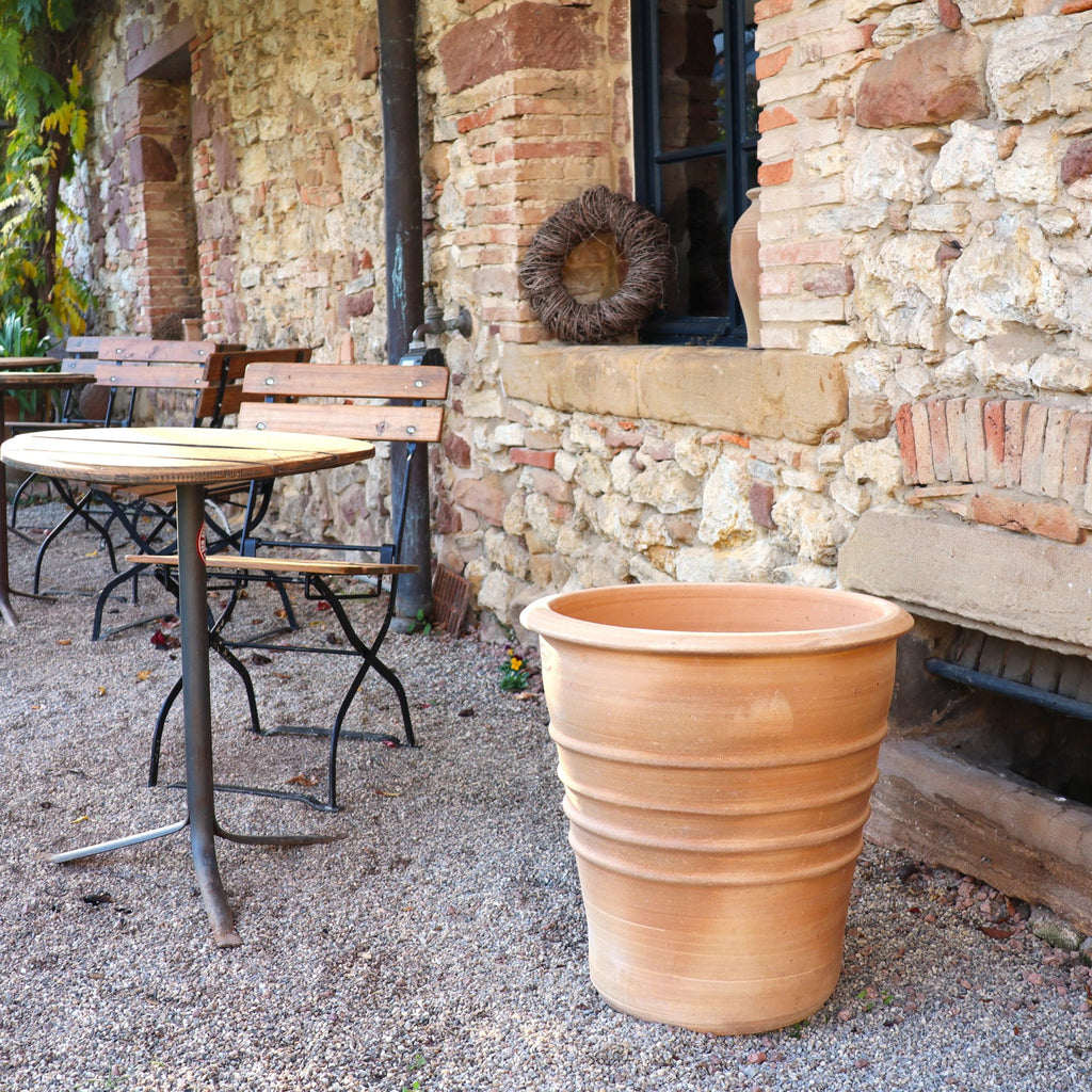 Ein Terracotta Blumentopf "Aster", H=50 cm, steht leer auf Kies neben einem runden Holztisch und Stühlen vor einem rustikalen Steinhaus mit kleinem Fenster und Zierkranz.