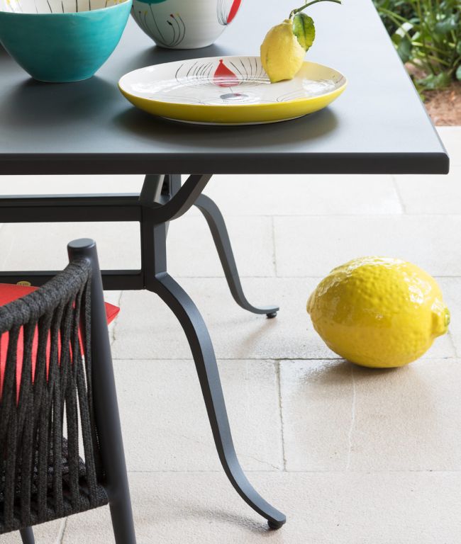 Ein quadratischer Tisch Toscana in Schwarz ist mit bunten Schalen und einem gelben Teller mit Zitronenmotiv geschmückt und ergibt ein elegantes Tableau. Unter dem Tisch ziert eine große dekorative gelbe Zitrone den Boden. Ein schwarzer Stuhl mit rotem Kissen ragt in den Vordergrund und verleiht Charme, der an die Toskana erinnert.