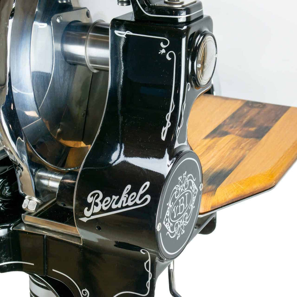 Eine Nahaufnahme der Berkel Volano B114 in Schwarz, einer manuellen Aufschnittmaschine mit stabilem Standfuß und angeschlossenem Fassholzbrett, das aufwendige weiße Verzierungen aufweist. Das runde Emblem mit der Prägung „Berkel“ ist deutlich zu erkennen.