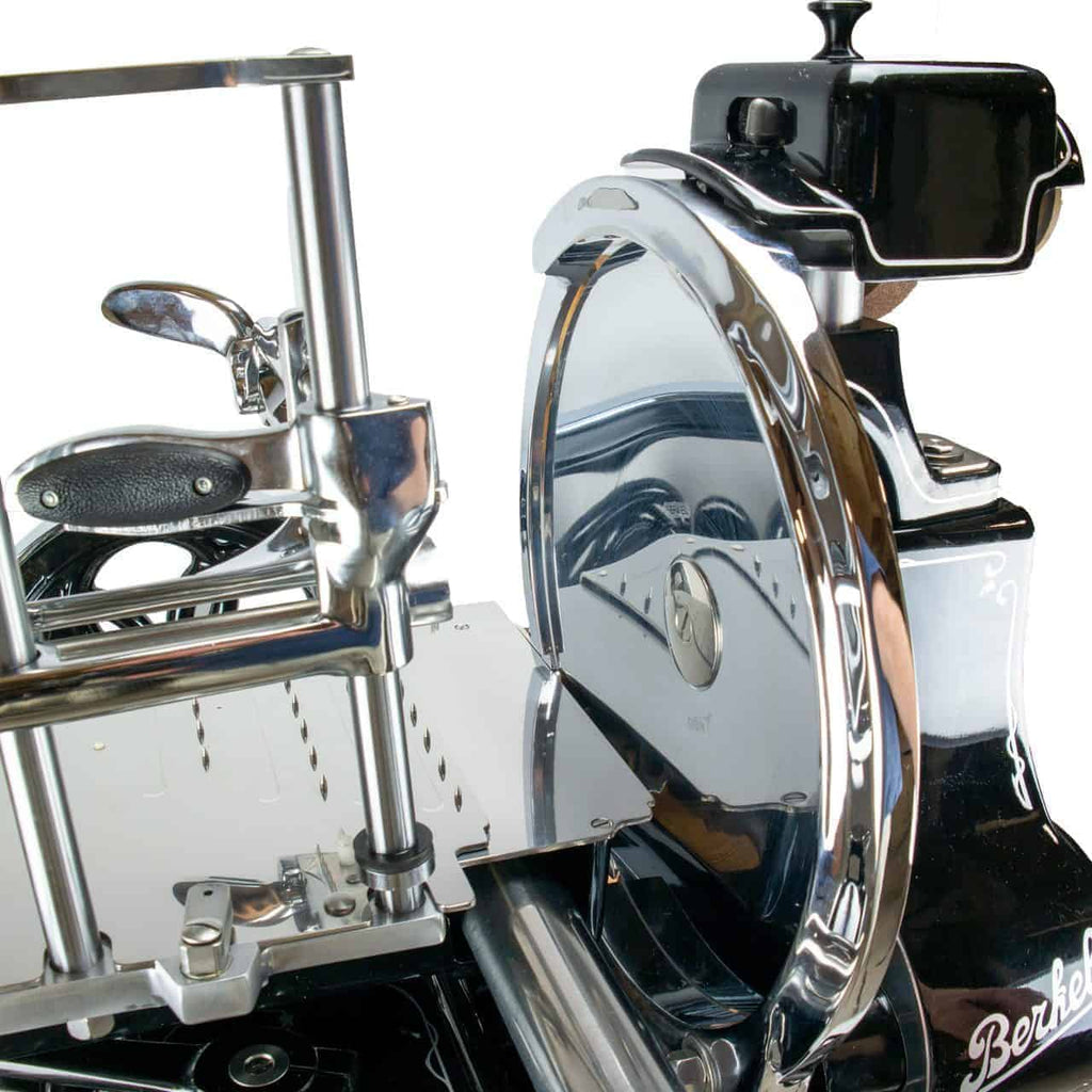 Eine antike Fleischschneidemaschine mit Handkurbel, glänzenden, verchromten Teilen und einem schwarzen Sockel. Die Maschine hat eine runde Schneide, einen Griff mit Griff und aufwendige Metalldetails. Der Markenname „Berkel“ ist auf der rechten Seite des Sockels zu sehen.