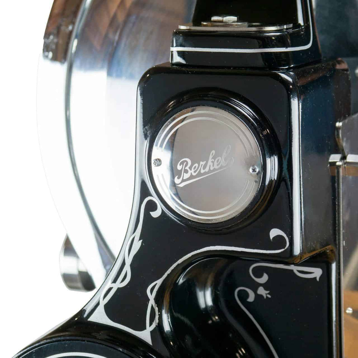 Nahaufnahme eines schwarzen Berkel-Allesschneiders im Vintage-Stil mit verzierten weißen Details. Der Fokus liegt auf dem Berkel-Logo, das gut sichtbar in der Mitte angebracht ist. Im Hintergrund ist ein Teil der Edelstahlklinge des Allesschneiders zu sehen.