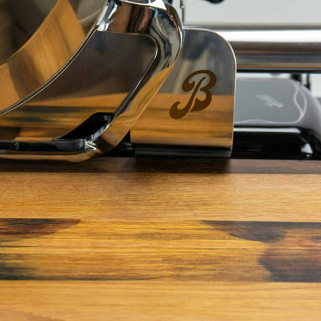 Eine Nahaufnahme eines glänzenden Fleischschneiders aus Metall mit einem in den Klingenschutz eingravierten „B“-Logo, der auf einer Holzoberfläche liegt. Die reflektierenden Oberflächen des Schneiders und die Holzmaserung des Schneidebretts sind deutlich sichtbar.