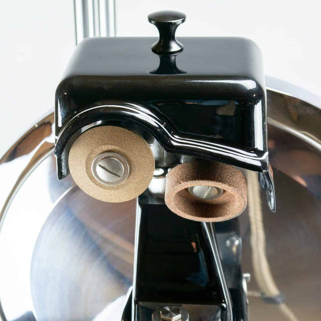 Nahaufnahme eines mechanischen Vintage-Geräts, das an die manuelle Aufschnittmaschine Berkel Volano B114 mit Ständer und Schneidebrett erinnert und zwei strukturierte zylindrische Komponenten aufweist – eine schwarze und eine beige – die an einer Metallstruktur befestigt sind. Das glänzende schwarze Gehäuse mit einem kleinen Knopf oben ergänzt die reflektierende Metalloberfläche dahinter und ruft den Charme einer manuellen Aufschnittmaschine hervor.