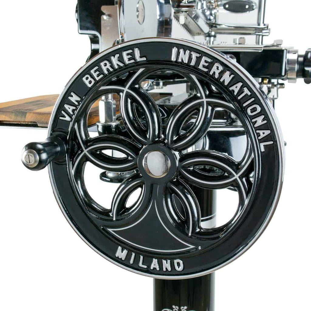 Nahaufnahme eines antiken Schwungrads aus schwarzem Metall auf einer Berkel Volano B114 manuellen Aufschnittmaschine mit Standfuß und Fassholzbrett, mit aufwendigen dekorativen Ausschnitten und dem Text „VAN BERKEL INTERNATIONAL MILANO“. Die metallischen Oberflächen sind glatt und reflektierend und heben die Handwerkskunst des Geräts hervor.