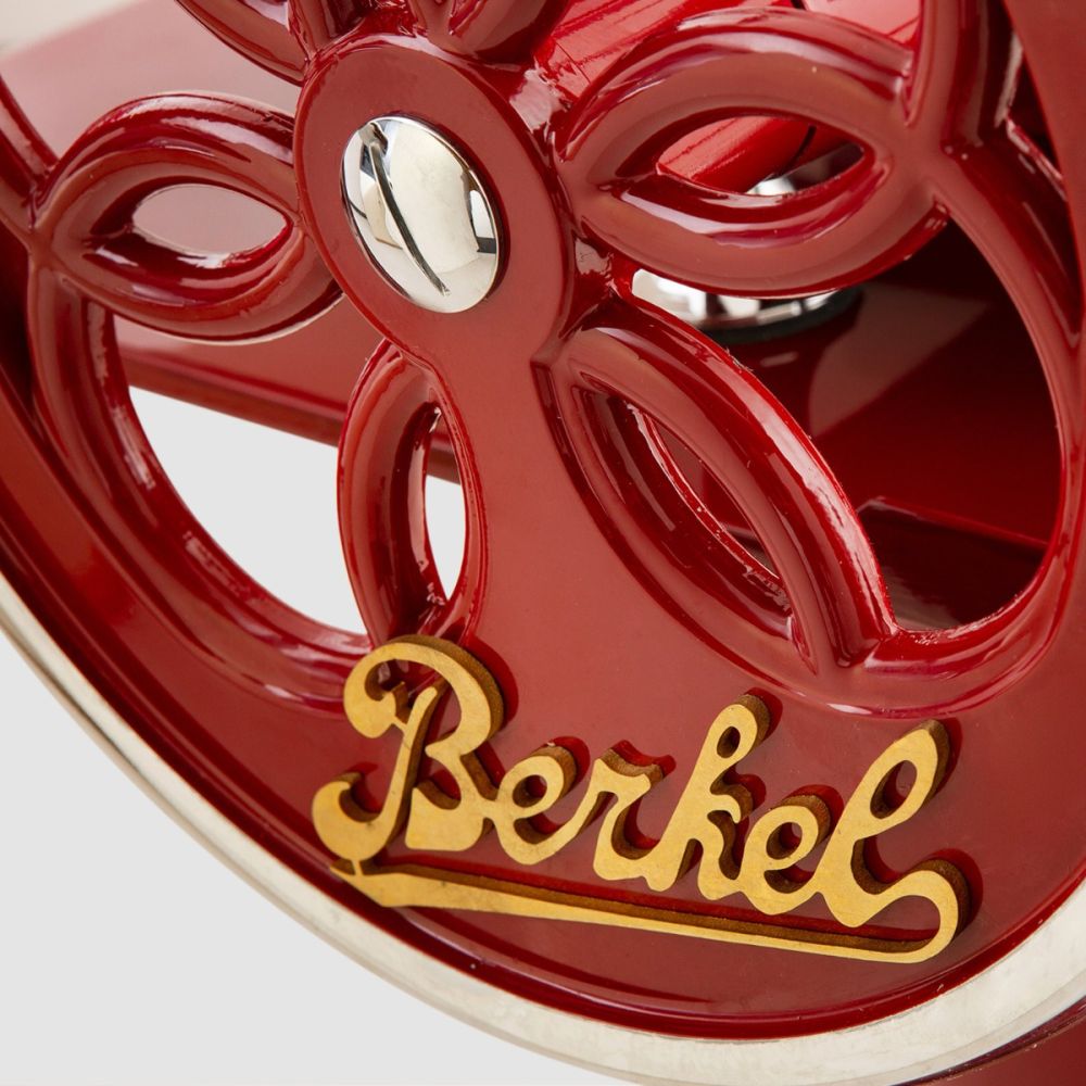 Nahaufnahme einer roten Berkel Volano B2 Schwungradmaschine im Vintage-Stil. Das Bild zeigt ein kompliziertes Blumenmuster auf dem Rad, wobei der Markenname „Berkel“ in goldener Schrift deutlich zu erkennen ist. Die Wunderschöne Aufschnittmaschine verfügt über einen polierten Metallknopf in der Mitte des Rads.