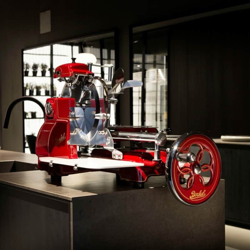 Eine glänzende rot-silberne Vintage-Fleischschneidemaschine von Berkel steht auf einer dunklen Arbeitsplatte in einer modernen Wohnküche. Die Maschine verfügt über komplizierte mechanische Teile und ein handgekurbeltes Rad, das einen Kontrast zum glatten, modernen Hintergrund bildet.