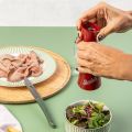 Eine Person mahlt Pfeffer mit der Berkel Pfeffer und Salz Mühle Rot über einer Schüssel mit Salat, neben einem Teller mit Wurstaufschnitt und einem Messerschärfer einfach, alles auf einem hellgrünen Tisch.
