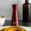 Eine rote "Berkel Pfeffer und Salz Mühle Rot" steht auf einer dunklen Fläche neben einer Schale mit Zitronen, einem Messerschärfer einfach und einer dunklen Glasflasche, alles vor einem hellgrauen Hintergrund.