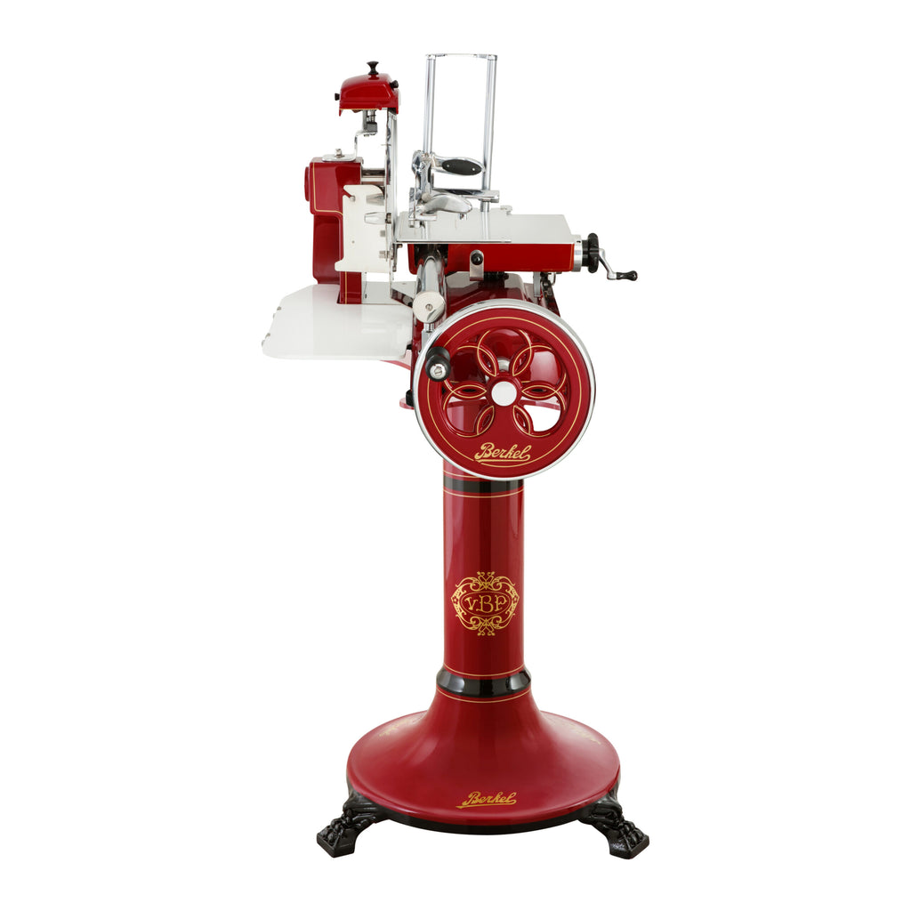 Die Berkel Volano B3 Rot Schwungradmaschine mit Standfuß und Fassholzeinlegebrett ist eine rot-weiße Fleischschneidemaschine im Vintage-Stil mit großer Handkurbel und aufwendigen Verzierungen und steht auf einem stabilen Sockel. Die Schneidemaschine verfügt über ein rundes Messer, einen Schutzbügel und eine Einfüllschale, alles in einem klassischen Design, das an Maschinen des frühen 20. Jahrhunderts erinnert.